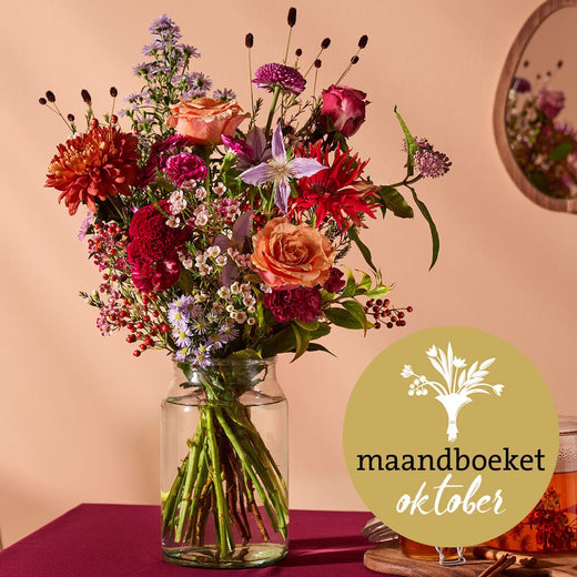 Maandboeket oktober met rozen, dahlia’s en herfstbloemen in een glazen vaas, op een sfeervolle tafel.