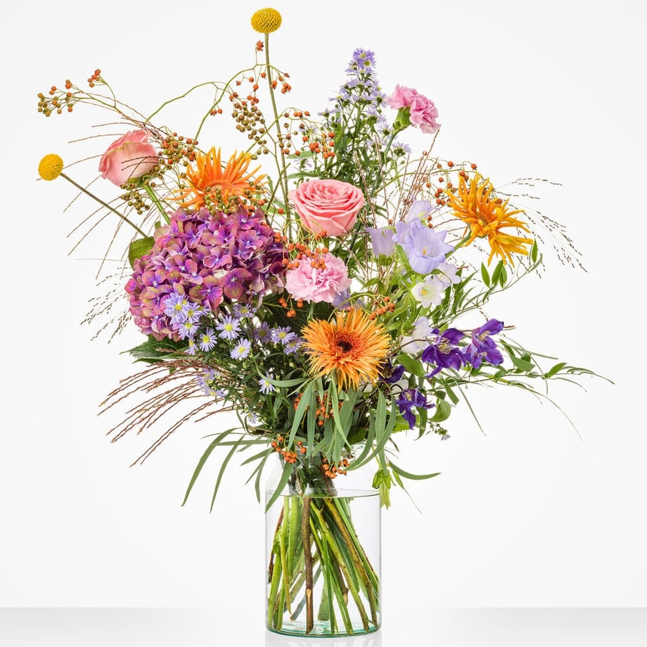 Groot bloemboeket in een glazen vaas met diverse kleurrijke bloemen, waaronder roze rozen, paarse hortensia’s, oranje gerbera’s, paarse campanula en groen loof.