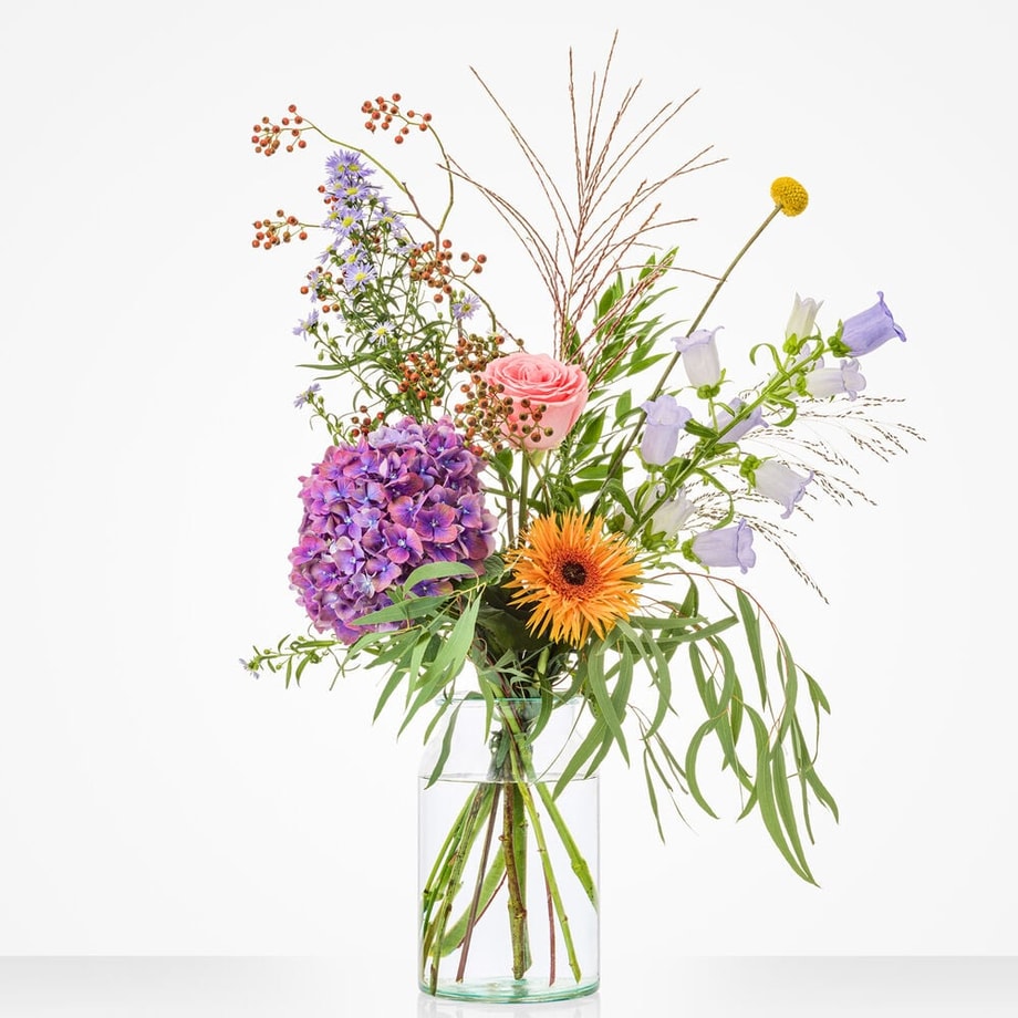 Boeket in glazen vaas met diverse bloemen, waaronder een roze roos, paarse hortensia, oranje gerbera, paarse campanula, en groene bladeren.