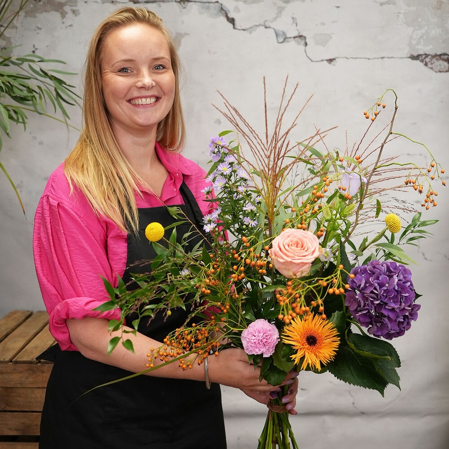 Vrouw in roze blouse en zwarte schort poseert lachend met een kleurrijk boeket in haar handen, bestaande uit bloemen zoals een roze roos, paarse hortensia, oranje gerbera en diverse groene takken.