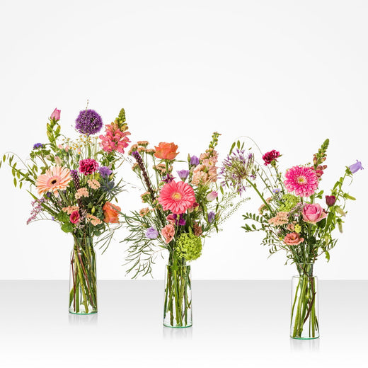 Drie kleurrijke veldboeketten met roze, paarse en oranje bloemen in glazen vazen – perfect voor moederdag