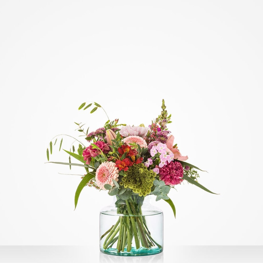 Klein boeket met roze en rode bloemen, waaronder gerbera’s, rozen en alstroemeria, in glazen vaas tegen witte achtergrond