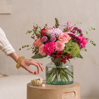 Boeket met roze en fuchsia bloemen, waaronder rozen, chrysanten en alstroemeria, in glazen vaas op tafel met hand die chocolaatje pakt