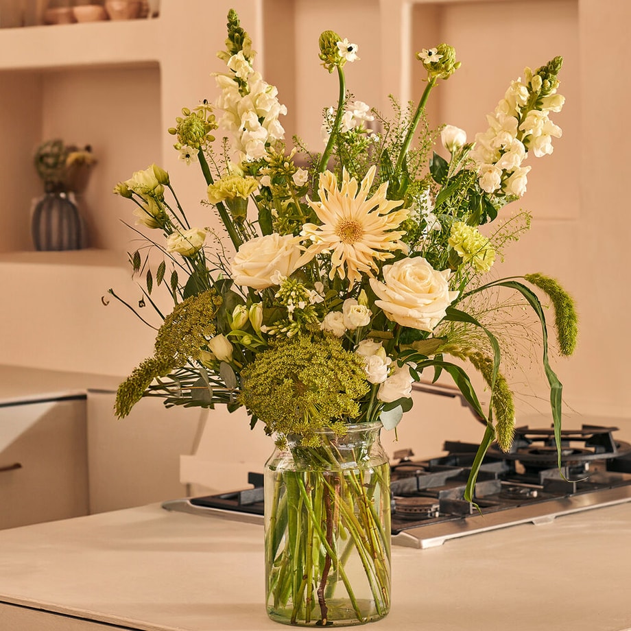 Mooi wit boeket met rozen, gerbera’s en lisianthus in glazen vaas op beige keukentafel