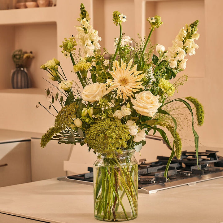 Mooi wit boeket met rozen, gerbera’s en lisianthus in glazen vaas op beige keukentafel