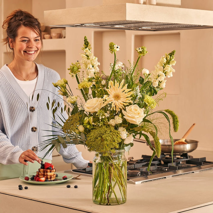 Vrouw in een keuken met een boeket van witte bloemen, waaronder witte rozen en gerbera's, in een glazen vaas op een aanrecht.