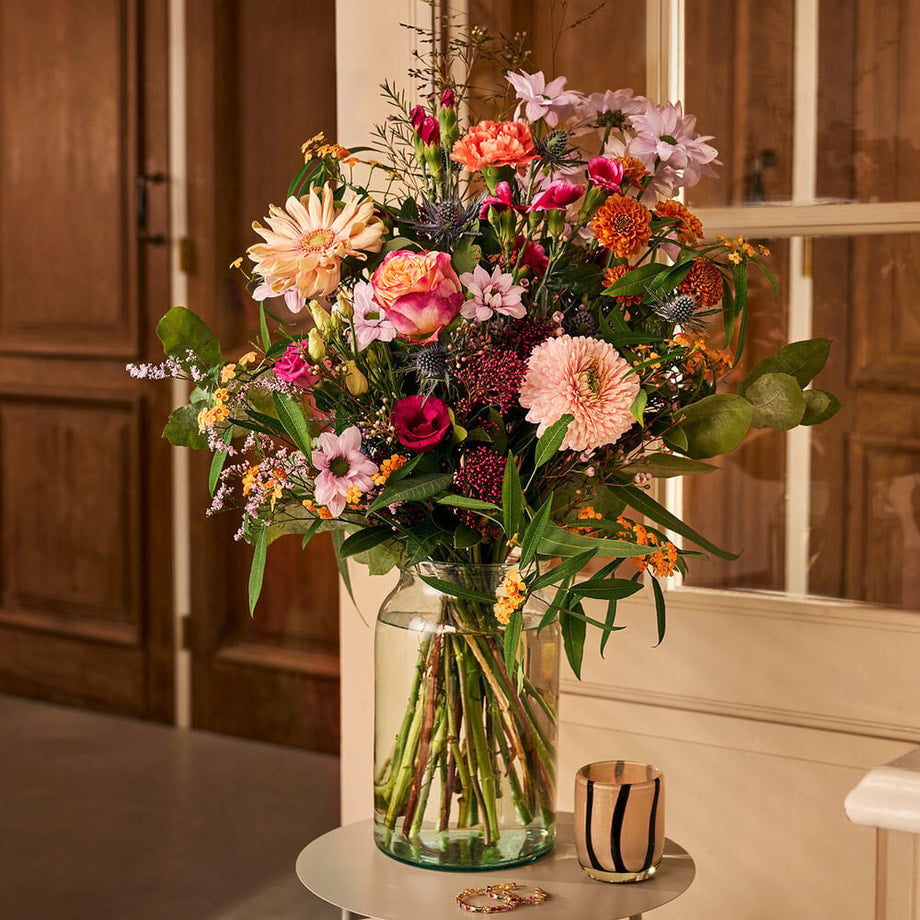 Sfeervol seizoensboeket met rozen, gerbera’s en seizoensbloemen in een glazen vaas in een gezellig interieur.