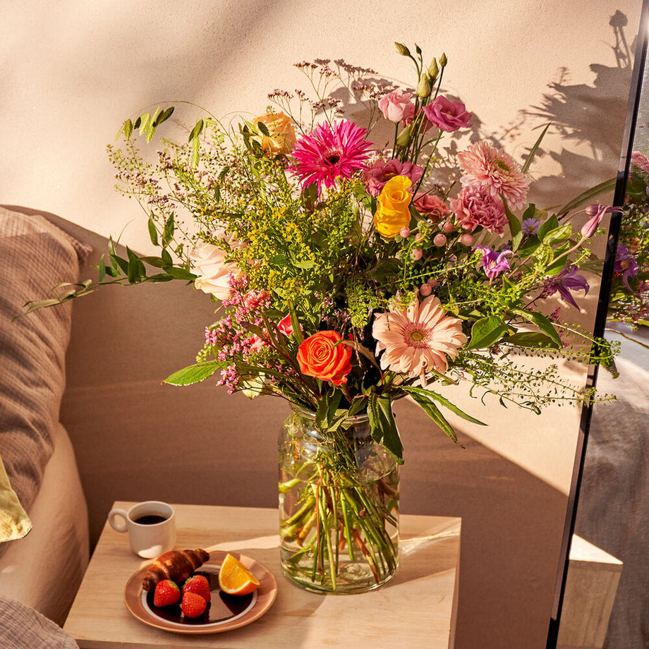 Sfeervolle setting met het opkikker boeket in een slaapkamer, met bloemen in vrolijke kleuren naast een ontbijtje met koffie en fruit