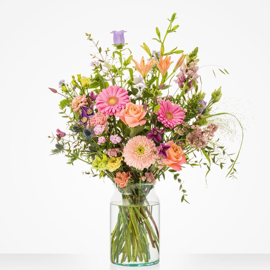Groot boeket met roze gerbera’s, rozen, paarse klokjesbloemen, oranje lelies en groen blad in een glazen vaas.