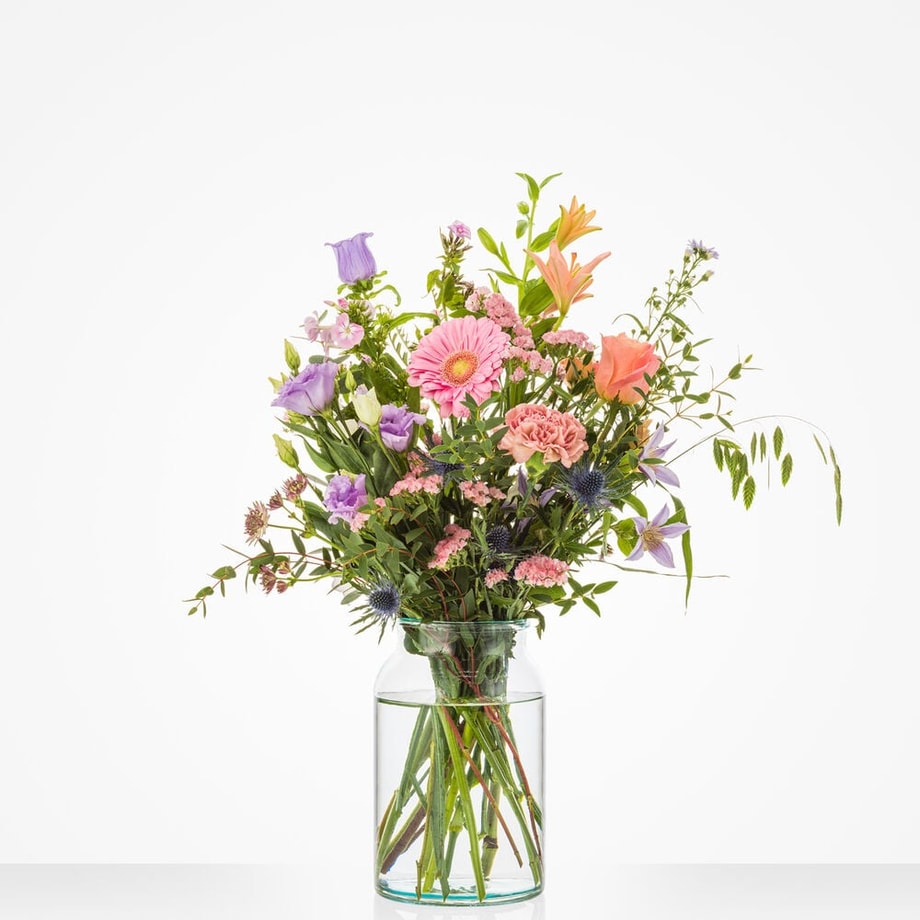 Klein boeket met roze gerbera’s, paarse lisianthus, oranje lelies en groen blad in een glazen vaas.