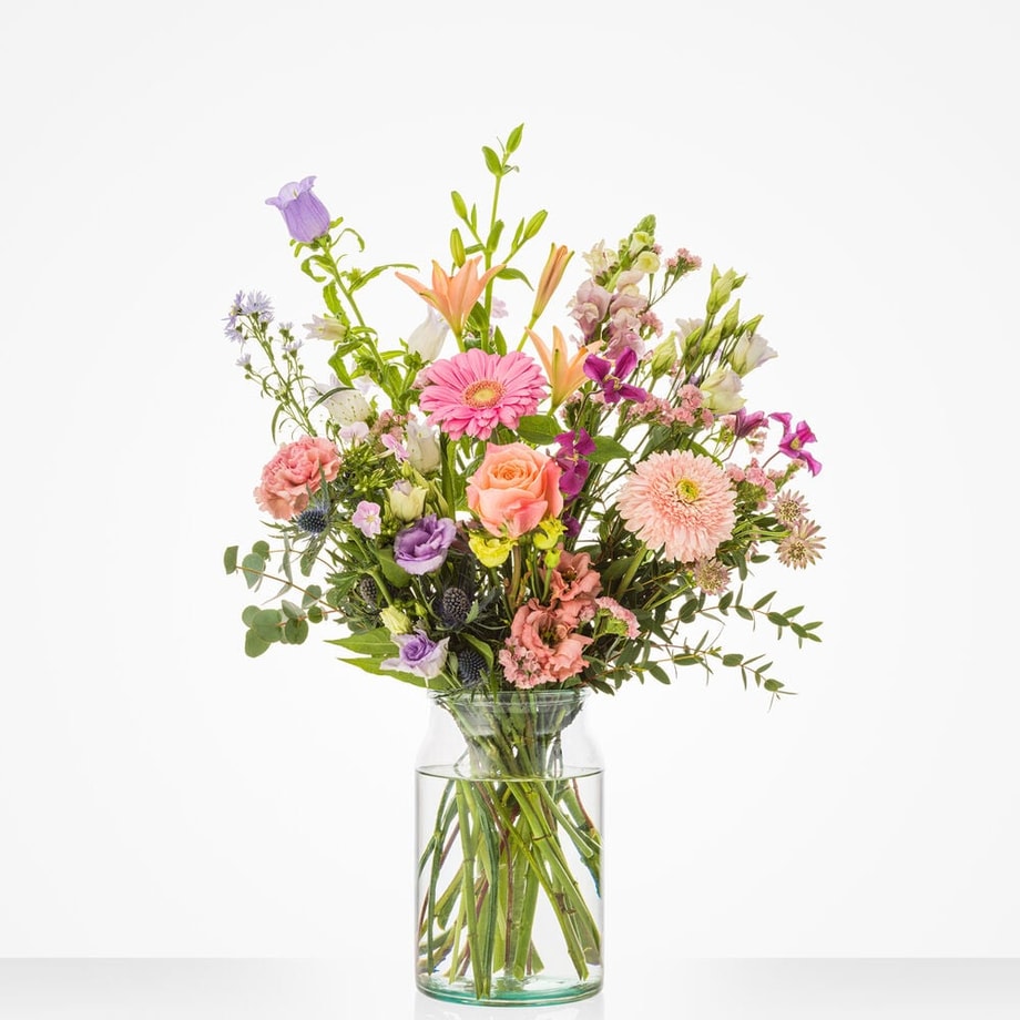 Middelgroot boeket met roze gerbera’s, rozen, paarse lisianthus, oranje lelies en groen blad in een glazen vaas.