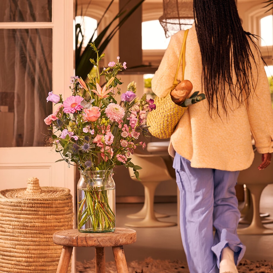 Boeket van pastel bloemen in een glazen vaas op een houten kruk, vrouw loopt weg met boodschappentas.
