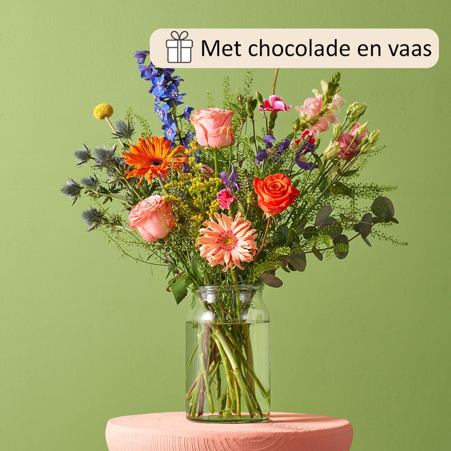 Sfeervol interieur met groene muur met een boeket in een glazen vaas op een roze tafeltje. Diverse bloemen zoals oranje rozen, roze gerbera’s, paarse lisianthus en blauwe ridderspoor.