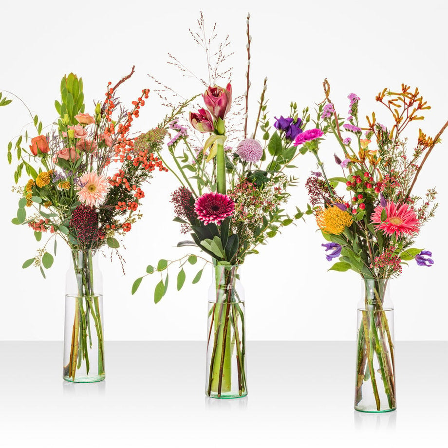 Drie kleurrijke winterboeketten met rozen, gerbera’s en seizoensbloemen in smalle glazen vazen.