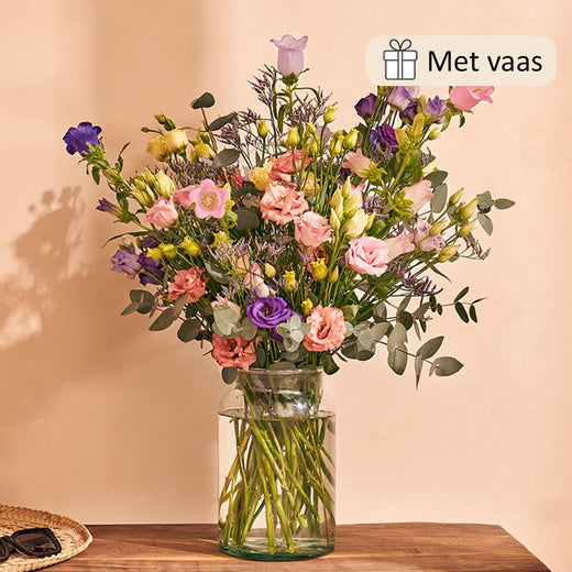 Stijlvolle interieursetting met een boeket in een glazen vaas, gevuld met kleurrijke bloemen zoals paarse lisianthus, roze rozen en eucalyptus bladeren, geplaatst op een houten tafel.