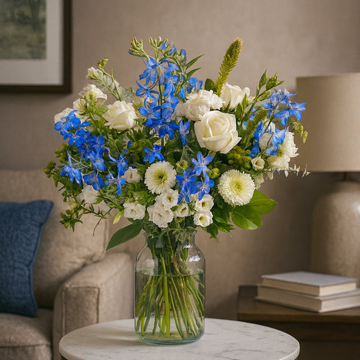 Sterkte boeket met witte en blauwe bloemen, waaronder rozen en delphinium, in glazen vaas op marmeren tafel in woonkamer