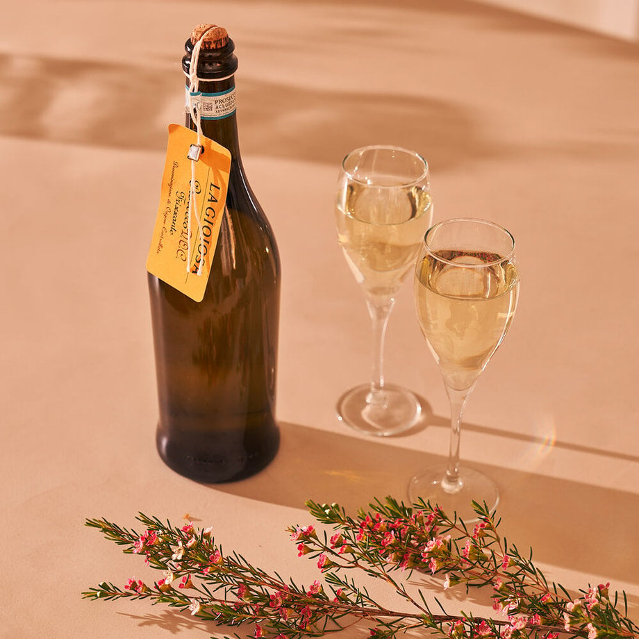 Een fles La Gioiosa prosecco en twee gevulde glazen op tafel, omringd door roze bloemen.
