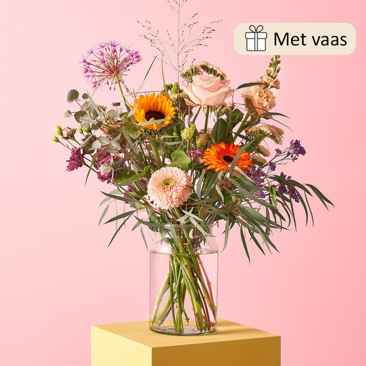 Boeket met zonnebloemen, gerbera’s, rozen, alliums, en eucalyptus in een glazen vaas, geplaatst op een geel blok tegen een roze achtergrond.