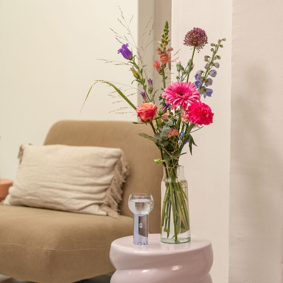 Vaas met bont boeketje van roze, paarse en oranje bloemen op bijzettafel naast beige fauteuil met kussen