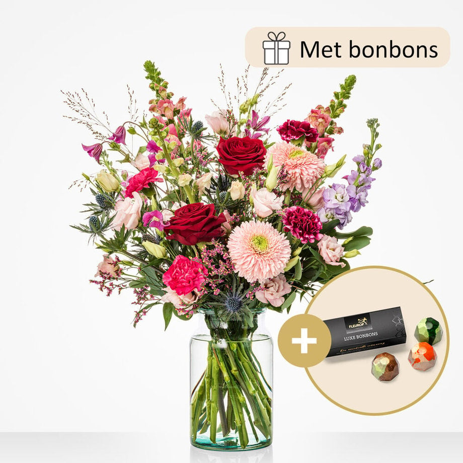 Groot boeket in een glazen vaas, vergezeld van luxe bonbons. Het boeket bevat bloemen zoals rode rozen, roze asters, lila leeuwenbek en paarse distels.
