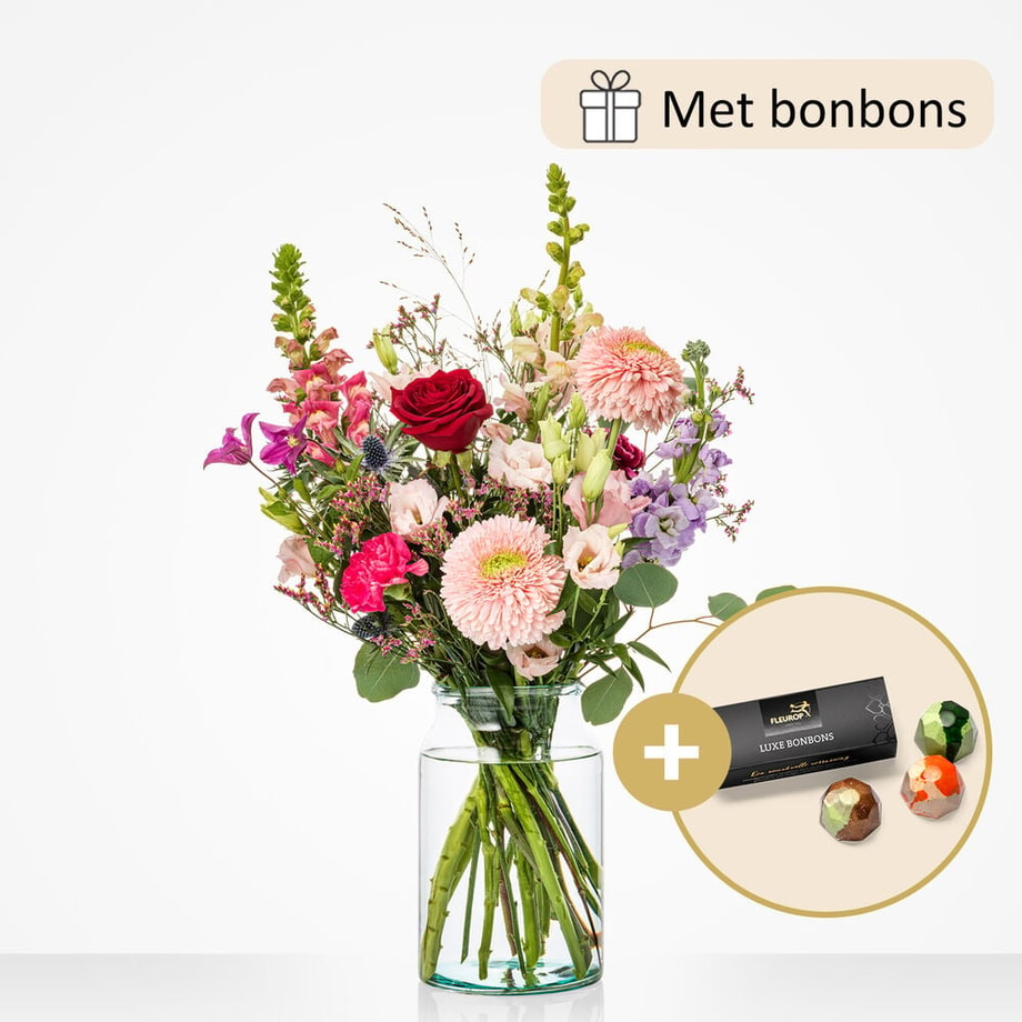 Een boeket in een glazen vaas met een bijpassende cadeauverpakking van luxe bonbons. Het boeket bevat bloemen zoals rode rozen, lichtroze gerbera’s, paarse distels en lila leeuwenbek.