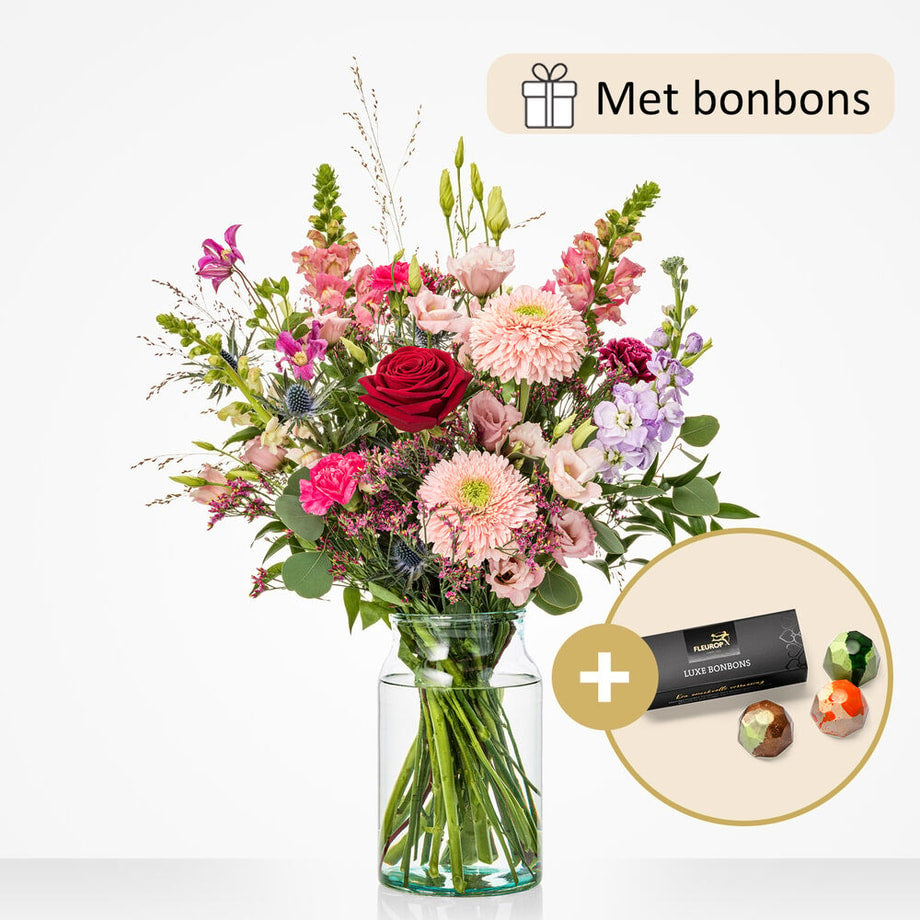 Een middelgroot boeket in een glazen vaas met luxe bonbons als cadeau. De bloemen in het boeket zijn onder andere rode rozen, lichtroze asters, paarse distels en lila leeuwenbek.
