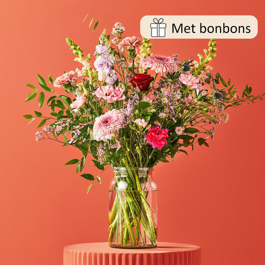 Een kleurrijk boeket in een glazen vaas, tegen een koraalrode achtergrond, met bloemen zoals roze gerbera’s, lila leeuwenbek, rode rozen en paarse distels.