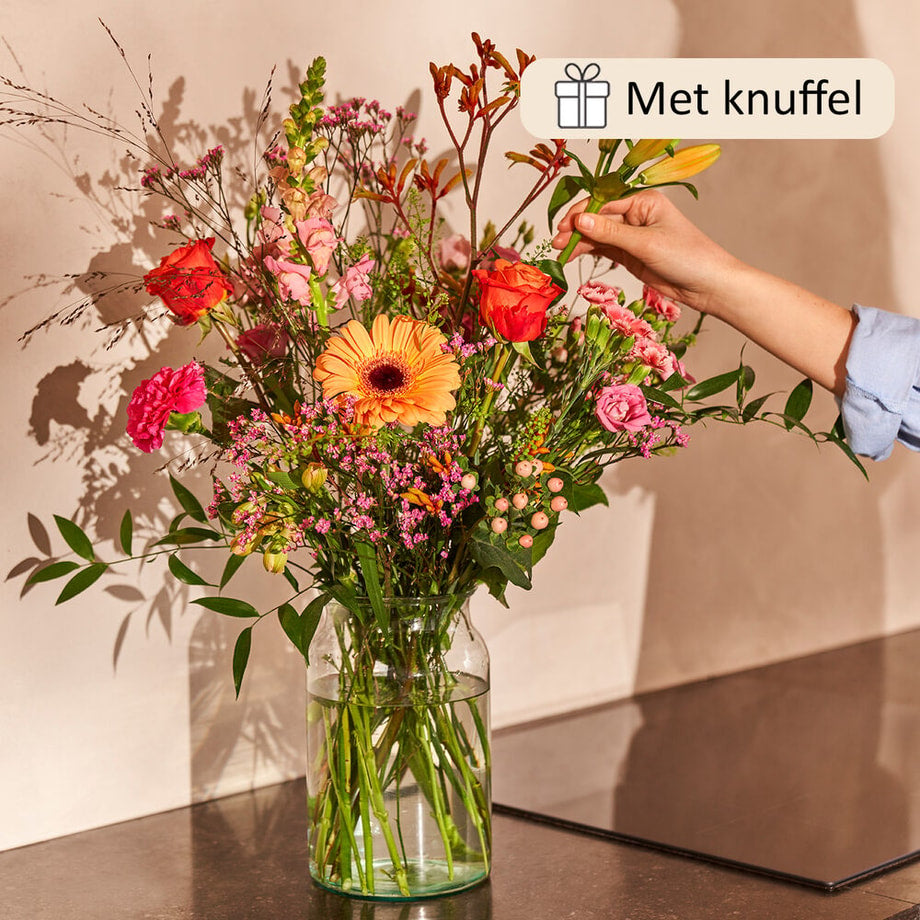 Boeket met een mix van bloemen zoals oranje gerbera’s, roze rozen, lila bloemen, en gele lelies in een glazen vaas. Een hand schikt bloemen in het boeket tegen een neutrale achtergrond, gepresenteerd met een knuffel.