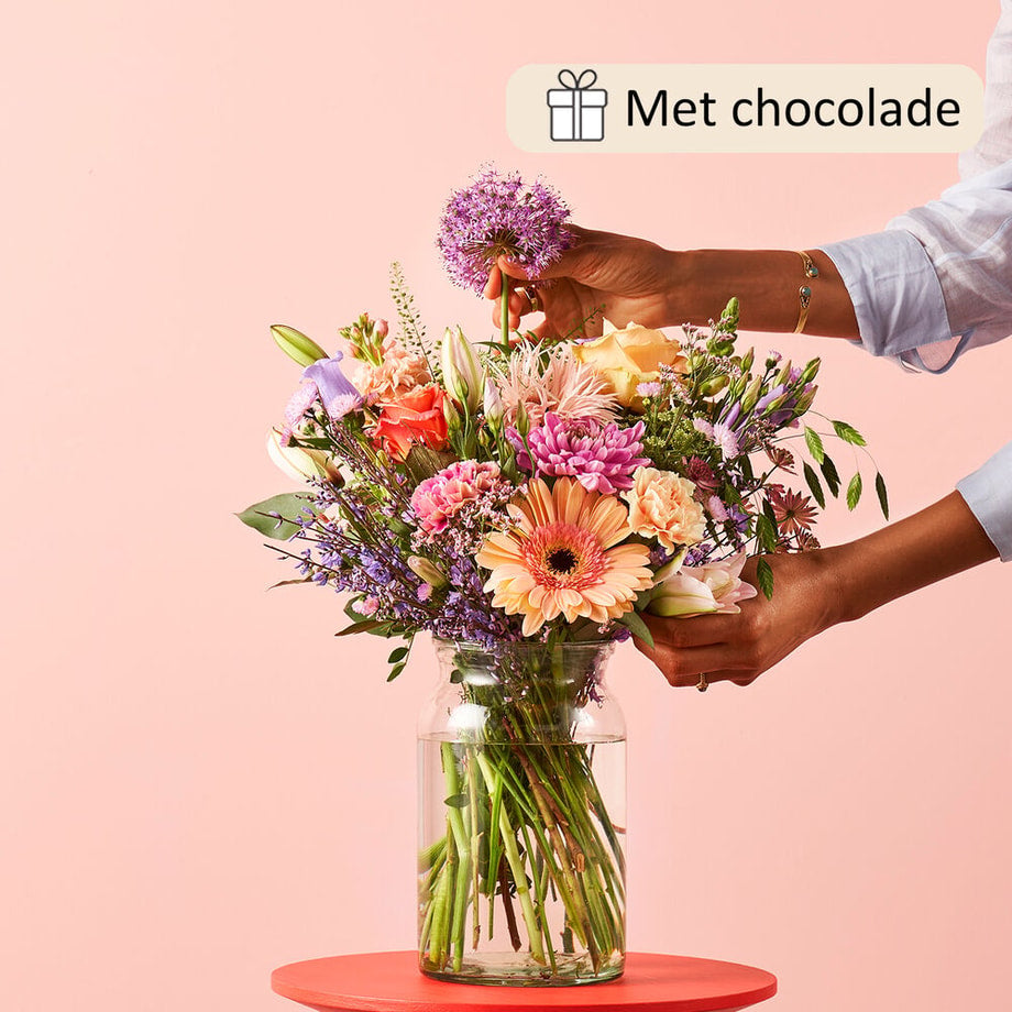 Boeket met een mix van bloemen zoals gerbera’s, rozen, lelies, en paarse bloemen in een glazen vaas. Een hand voegt een bloem toe aan het boeket tegen een roze achtergrond, gepresenteerd met luxe chocolade.