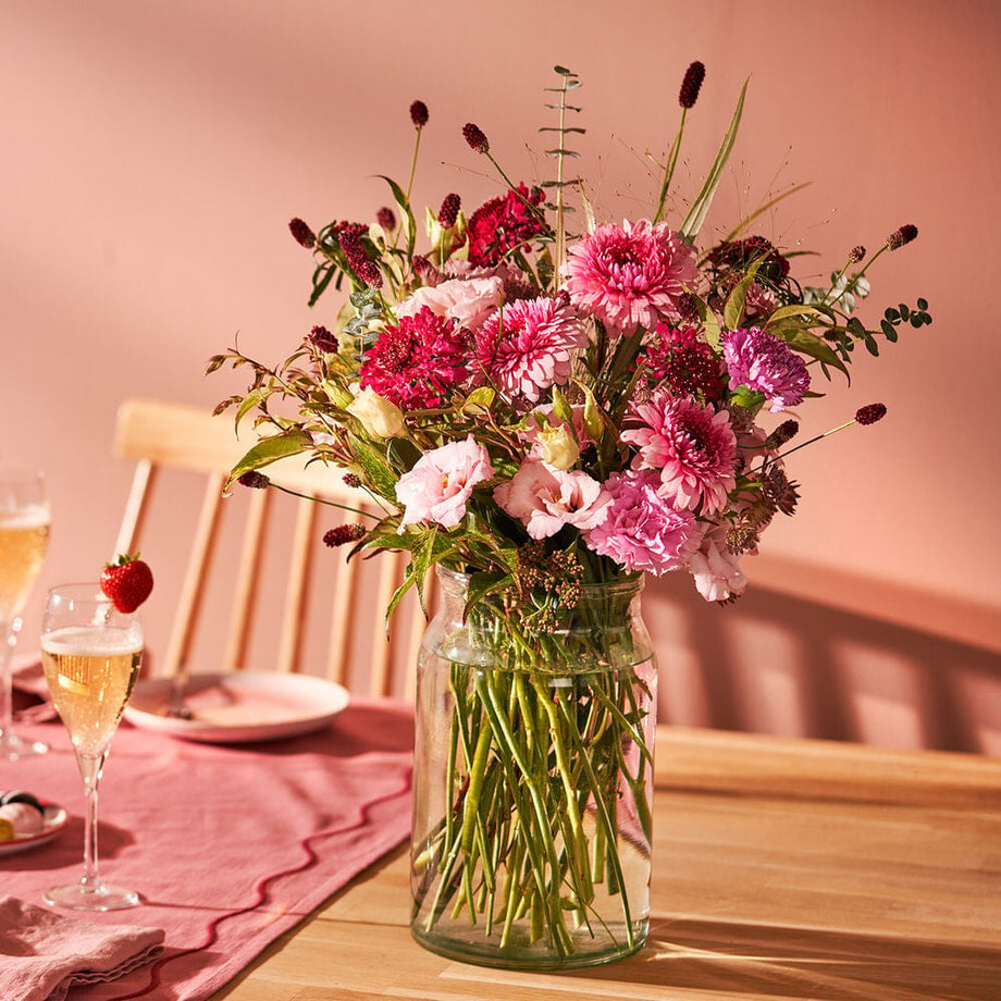 Boeket voor Moederdag met roze en paarse bloemen in een glazen vaas, op een houten tafel met roze servies en champagneglazen
