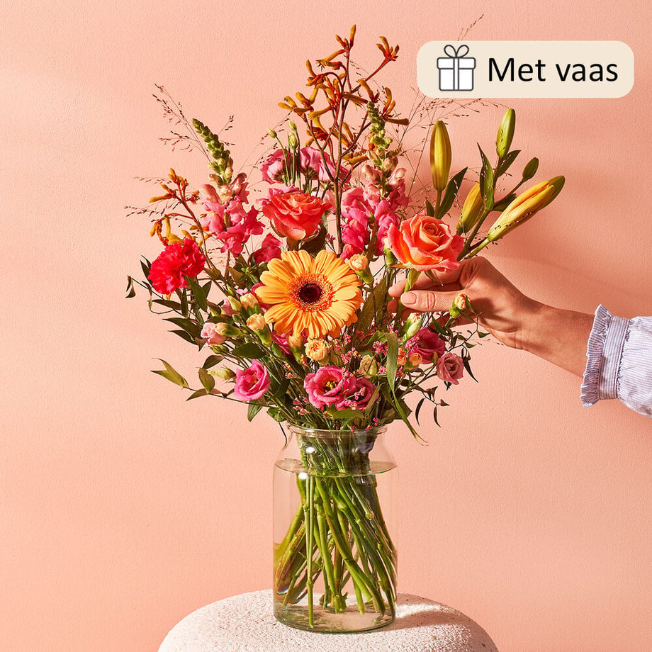 Boeket met een mix van bloemen zoals roze rozen, oranje gerbera’s, en gele lelies in een glazen vaas tegen een roze achtergrond. Een hand reikt naar het boeket.