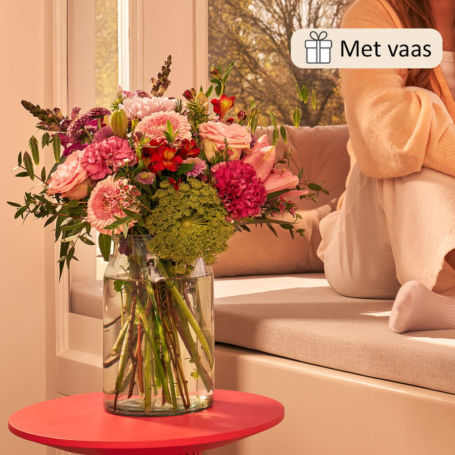 Boeket in een glazen vaas met een mix van bloemen zoals roze rozen, rode alstroemeria, groene bolvormige bloemen, en paarse anjers, geplaatst op een tafel bij een raam, met een warme, huiselijke sfeer.