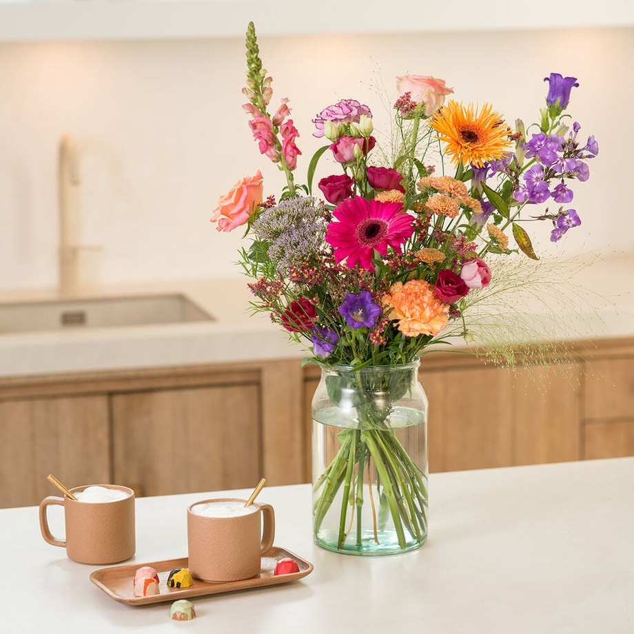 Kleurrijk veldboeket met felle zomerbloemen in vaas op keukentafel naast koffiekopjes.