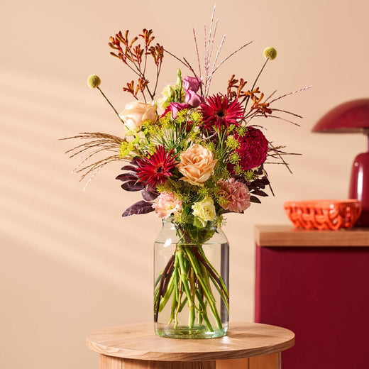 Herfstboeket met warme kleuren zoals rood, oranje en roze bloemen in een glazen vaas, geplaatst op een houten tafeltje.
