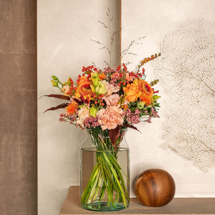 Zacht najaarsboeket met oranje rozen, rode bessen en roze bloemen in een glazen vaas.