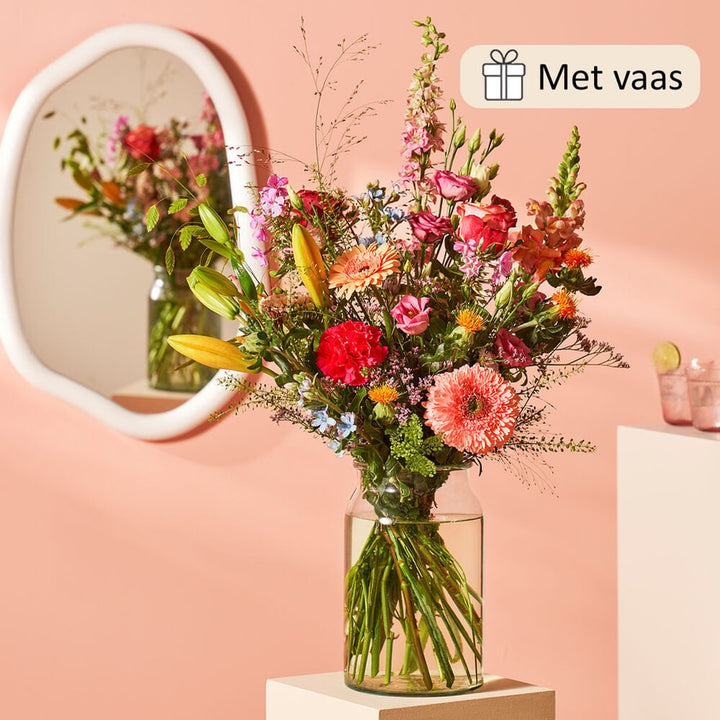 Boeket in een glazen vaas met een mix van zomerse bloemen zoals gele lelies, roze rozen, gerbera’s, leeuwenbekjes en rode anjers, geplaatst voor een roze achtergrond met een spiegel die het boeket reflecteert.