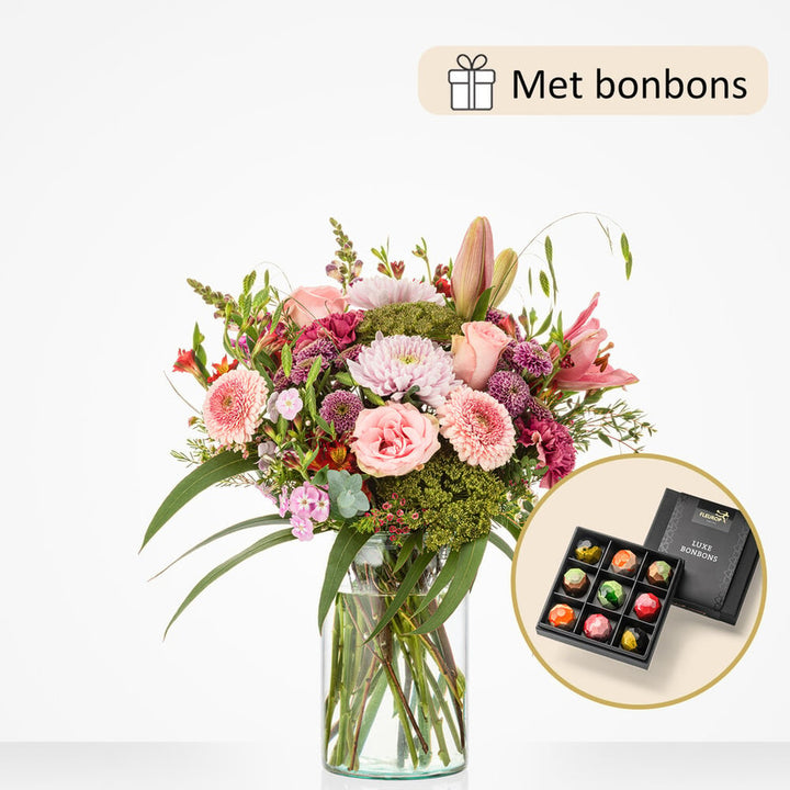 Groot boeket met bloemen zoals roze rozen, roze gerbera’s, groene bolvormige bloemen, paarse anjers en lelies, in een grote glazen vaas, geleverd met een luxe doos bonbons.