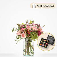 Middelgroot boeket met bloemen zoals roze rozen, alstroemeria, groene bolvormige bloemen, gerbera’s en paarse anjers, gepresenteerd in een glazen vaas met luxe bonbons als cadeau.