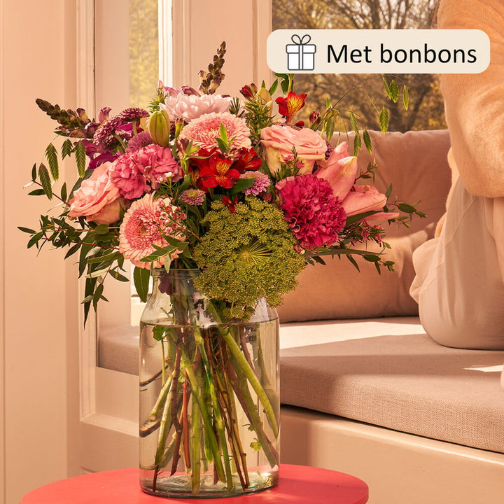 Boeket met een mix van bloemen zoals roze rozen, rode alstroemeria, groene bolvormige bloemen en paarse anjers, in een glazen vaas, geplaatst op een tafel bij een raam, geleverd met bonbons.