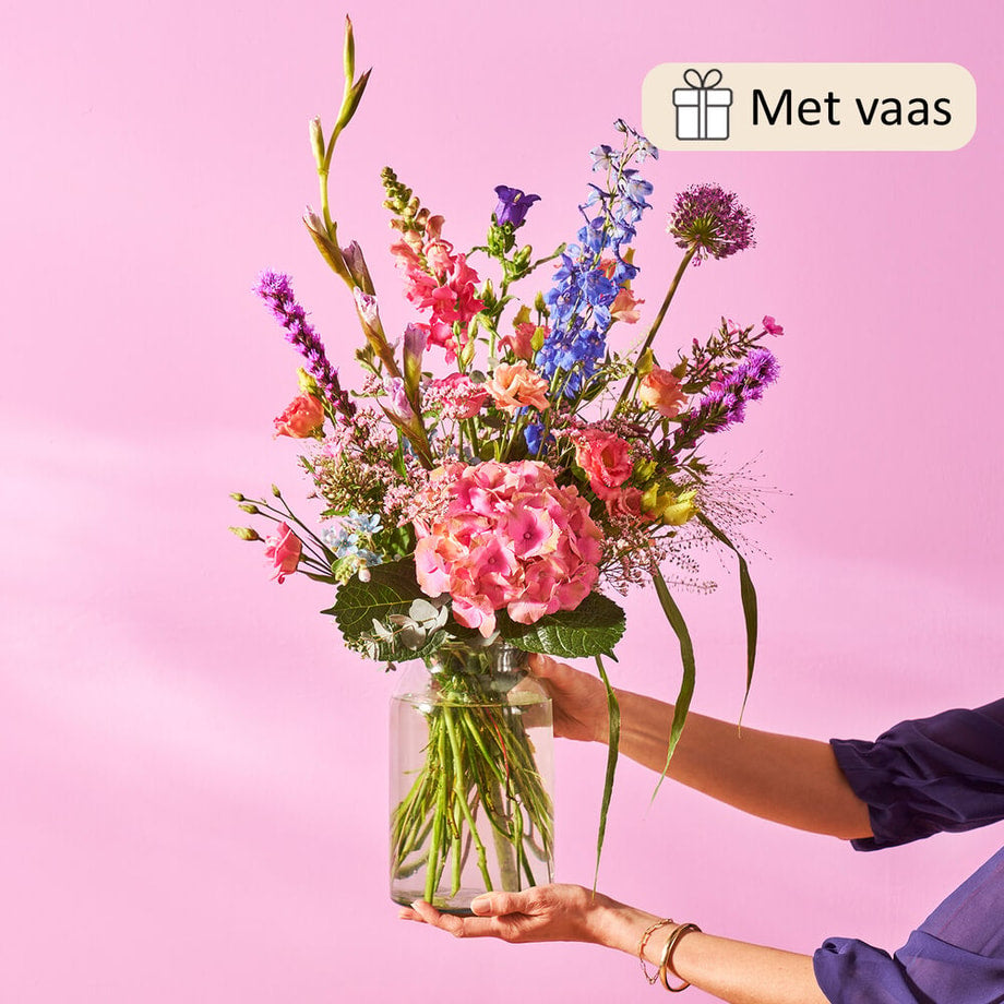 Sfeerfoto van een boeket met een mix van roze hortensia’s, blauwe riddersporen, leeuwenbekjes en andere veldbloemen, in een glazen vaas tegen een roze achtergrond, vastgehouden door een persoon.