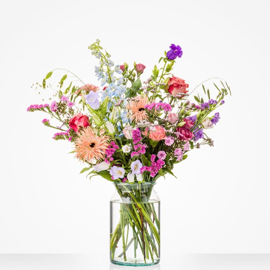 Groot boeket in een glazen vaas met een diverse mix van bloemen, waaronder roze gerbera’s, blauwe bloemen en rode rozen.