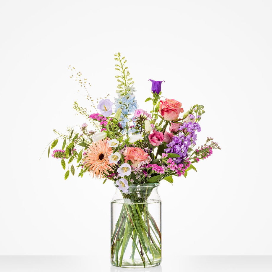 Klein boeket in een glazen vaas met een diverse mix van bloemen, waaronder roze gerbera’s, blauwe bloemen en rode rozen.