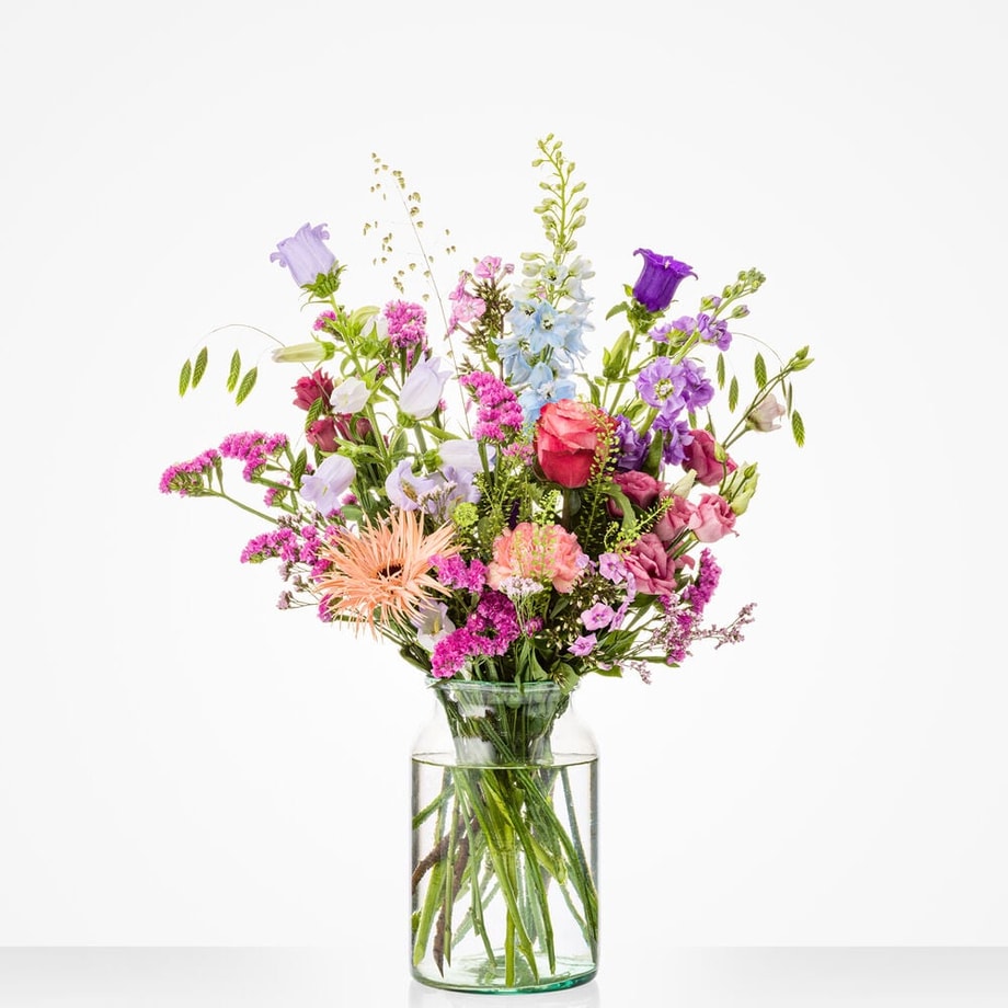 Middelgroot boeket in een glazen vaas met een diverse mix van bloemen, waaronder roze gerbera’s, blauwe bloemen en rode rozen.