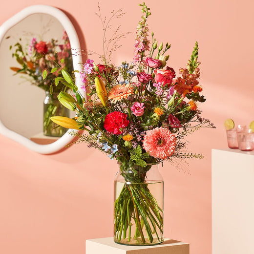 Groot boeket met een mix van kleurrijke bloemen in een glazen vaas op een witte sokkel tegen een roze muur met een spiegel op de achtergrond.