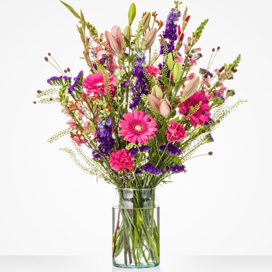 Groot boeket in een glazen vaas met een mix van kleurrijke bloemen, waaronder roze gerbera’s, lelies en paarse bloemen.