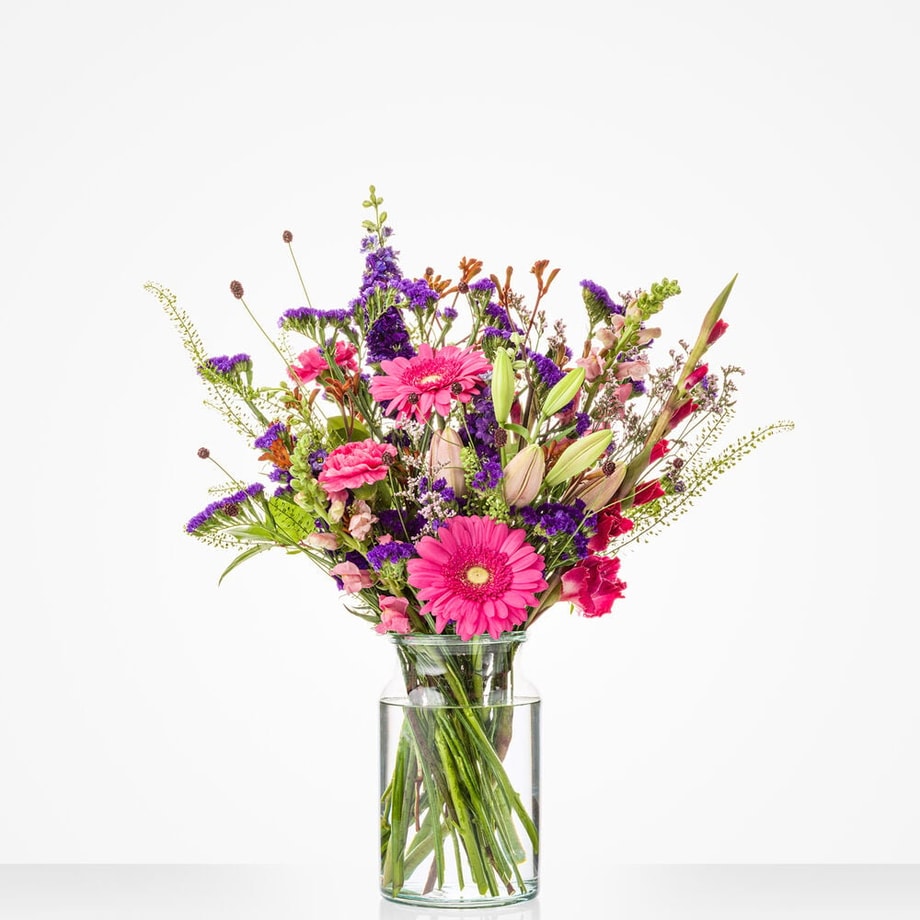 Klein boeket in een glazen vaas met een mix van kleurrijke bloemen, waaronder roze gerbera’s, lelies en paarse bloemen.