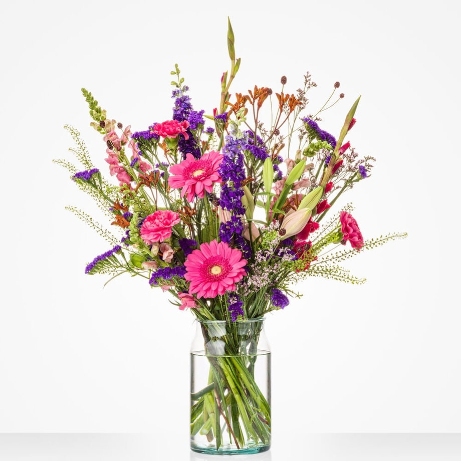 Middelgroot boeket in een glazen vaas met een mix van kleurrijke bloemen, waaronder roze gerbera’s, lelies en paarse bloemen.