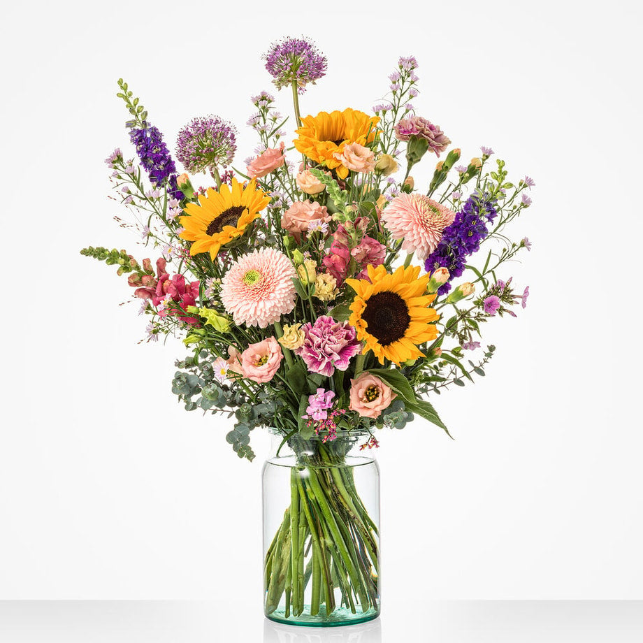 Groot boeket in een glazen vaas met een diverse mix van bloemen, waaronder zonnebloemen, roze en paarse bloemen.