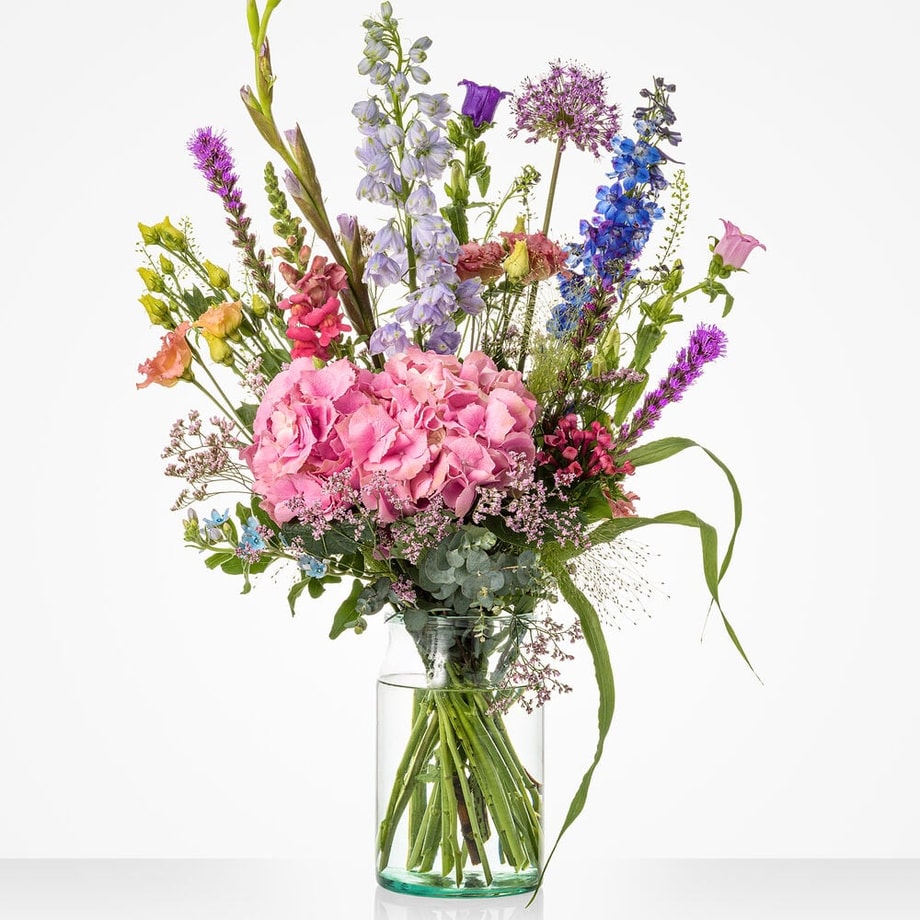 Groot gekleurd boeket in een glazen vaas met diverse bloemen, waaronder roze hortensia’s blauwe en paarse bloemen.