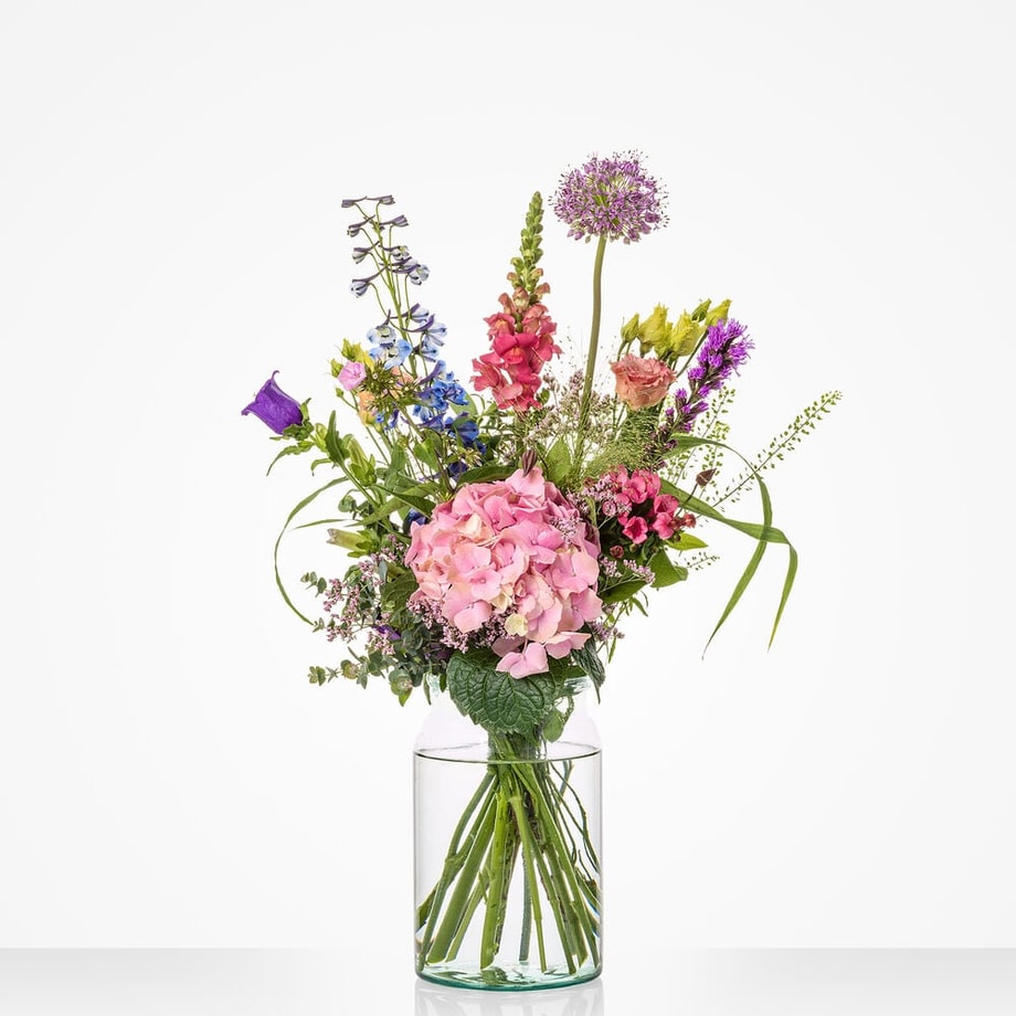 Klein gekleurd boeket in een glazen vaas met diverse bloemen, waaronder roze hortensia’s, blauwe en paarse bloemen.